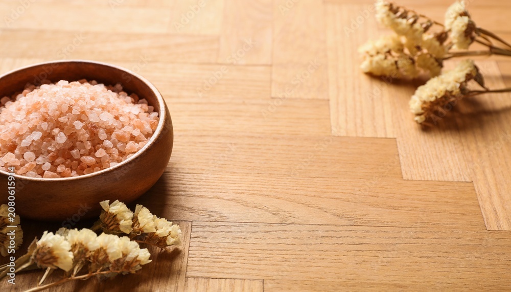 Fototapeta premium A wooden bowl filled with pink sea salt rests on a textured wooden surface, accompanied by dried flowers