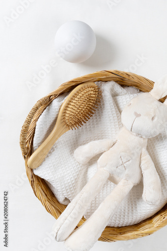 Flat lay of baby care product mockup background with plush bunny toy, wooden brush, and white towel in wicker basket. Top view with green leaves and copy space for organic newborn skincare brand.