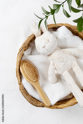 Flat lay of baby care product mockup background with plush bunny toy, wooden brush, and white towel in wicker basket. Top view with green leaves and copy space for organic newborn skincare brand.