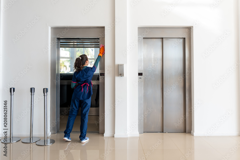 Fototapeta premium Female Janitor in Uniform Wiping Elevator Doors in Modern Building Lobby
