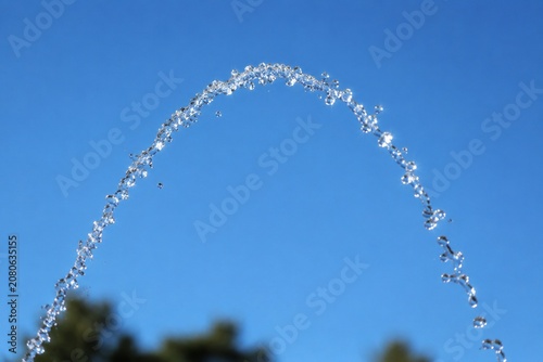 Graceful parabolic arc of a water fountain jet against a clear blue sky with glinting sunlight.