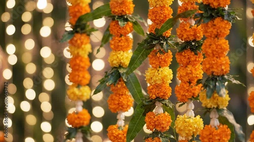 Festive orange marigold garlands hanging with green leaves against warm bokeh lights at an indian wedding.