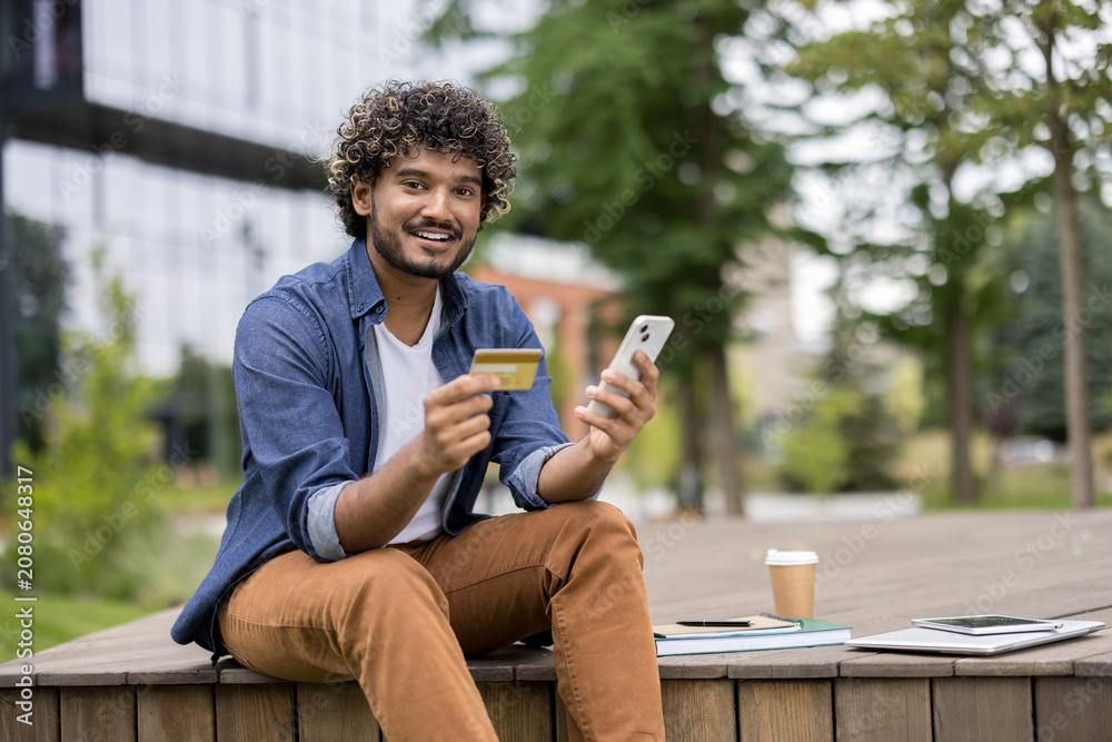 Fototapeta premium Young man with curly hair smiling while holding a credit card and smartphone, sitting outdoors, engaging in mobile banking, e-commerce, and digital finance transactions