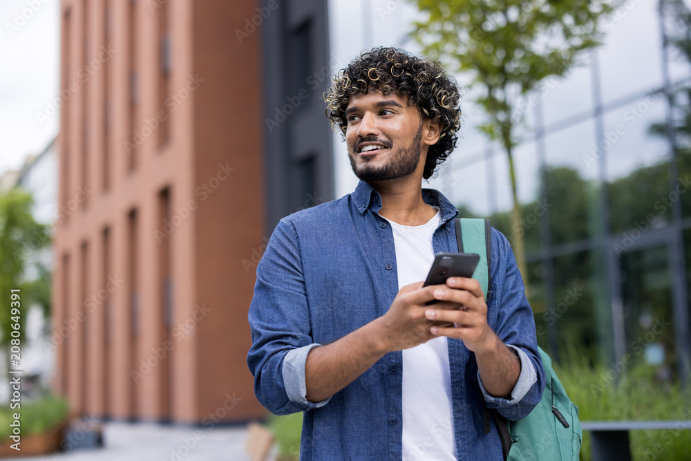 Obraz premium Young man with curly hair and backpack walking on a college campus while looking away and smiling, using his mobile phone for communication and connection