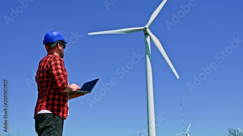 A windmill engineer stands outdoors in front of a wind turbine using a laptop. The blue sky is clear and the sun is shining brightly on the scene.
