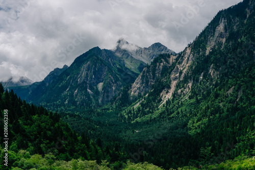 Dramatic mountain range shrouded in clouds, framed by lush green forests and valleys. A moody, expansive landscape.