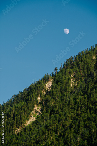 A crescent moon hangs in a clear blue sky above a dark, forested mountain range.