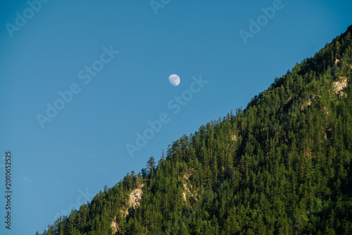 A crescent moon hangs in a clear blue sky above a dark green mountain range, silhouetted against the horizon.