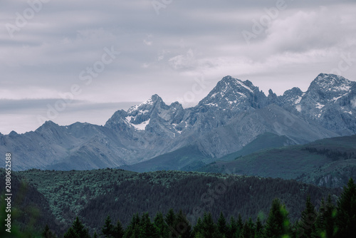 Dramatic mountain range under a cloudy sky, with a forested valley in the foreground.