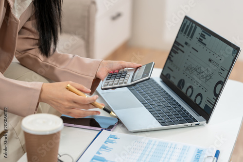 Businesswoman reviewing charts on a laptop while using a calculator and phone at a desk with financial papers and coffee, showing budgeting, analysis and office work.