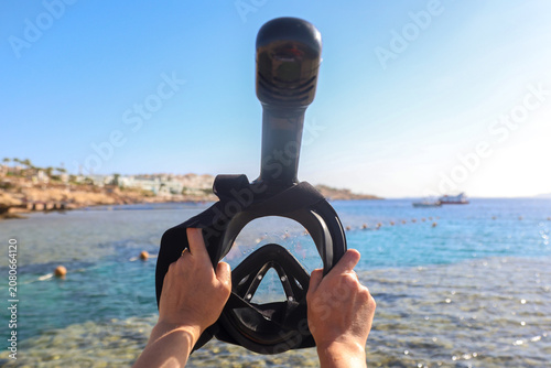 Woman holding snorkeling mask up against the bright blue sky with the sea in the background.