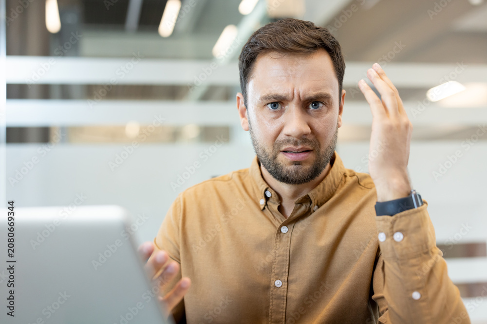 Fototapeta premium Frustrated man expressing confusion and annoyance while working on his laptop in a modern office, encountering a stressful business issue or technical problem
