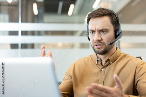 Male customer support agent with a headset responding to a challenging call, showing feelings of anger and stress while working on a laptop in a modern office setup
