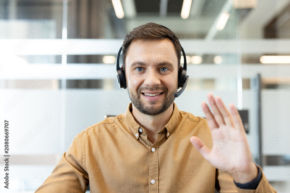 Obraz premium Man wearing a headset with a microphone, smiling confidently and waving hand while communicating during a remote business video call from an office