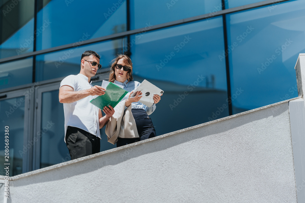 © qunica.com - Two business associates review documents and a tablet while standing on an office balcony. They discuss paperwork and digital files outside a modern glass building.