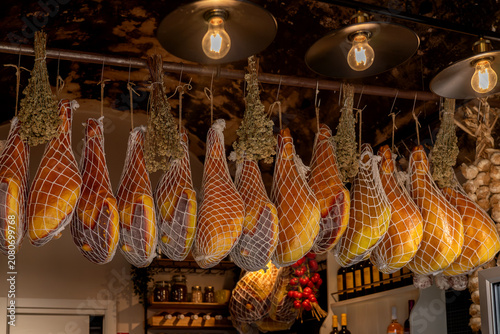 Canvastavla Traditional Italian cured hams hanging in a delicatessen with dried herbs and warm lighting