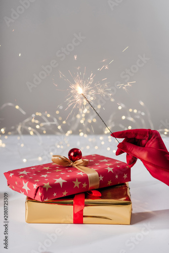 A hand in a red satin glove holding a burning sparkler over Christmas gift boxes, with a festive bokeh light background
