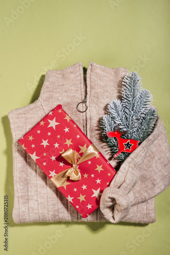Christmas gift concept with a folded beige knitted sweater, red star-patterned gift box, and a snowy pine branch with a deer ornament on a green background