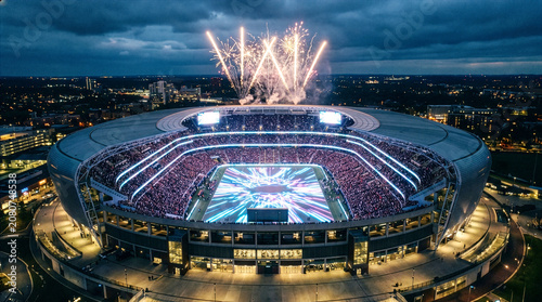 Aerial view of a modern sports stadium packed with fans at night, illuminated field, and spectacular fireworks exploding above