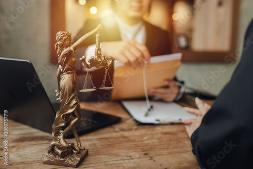 Fotografie The lawyer sat in front of a laptop in a room filled with documents and smartphones, with a hammer and scales beside him, advising his female client on matters of justice and legal principles