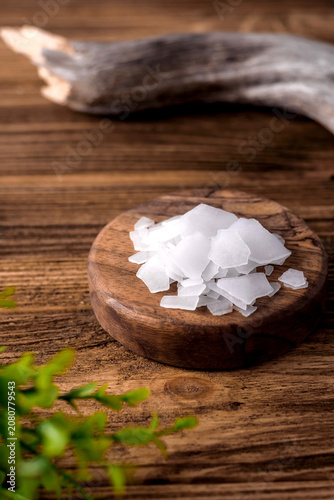 Natural bath salt flakes in a round wooden plate on a rustic wooden background with green leaves, spa and wellness concept