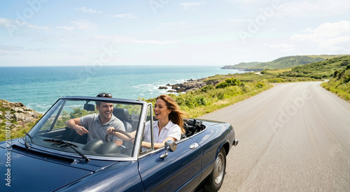 Happy couple driving a blue convertible car on a coastal road. Man and woman enjoying a summer road trip along the ocean. Vacation and travel lifestyle concept