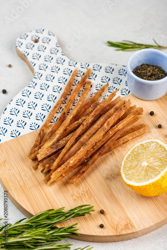 Dried fish sticks snack served on a round wooden cutting board with sea salt crystals, bright background