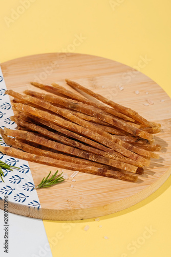 Dried fish sticks snack served on a round wooden cutting board with sea salt crystals, bright background