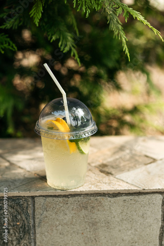 Refreshing summer lemonade with lemon and lime slices in a plastic takeaway cup with a straw, outdoors background