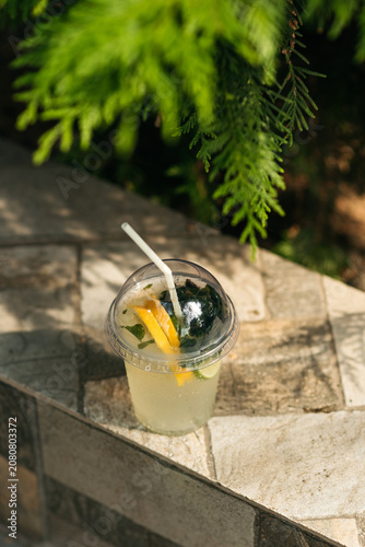 Refreshing summer lemonade with lemon and lime slices in a plastic takeaway cup with a straw, outdoors background