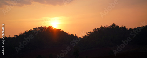 Golden sunset over forested mountain silhouettes beneath a warm orange sky, creating a peaceful natural landscape. Ideal for travel, nature backgrounds, outdoor scenery.