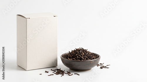 A simple presentation of loose leaf black tea in a dark ceramic bowl next to a plain white box on a white background