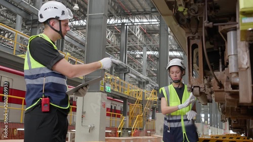 Two railway engineers meticulously inspect train undercarriage during detailed maintenance inspection within large industrial factory setting featuring machinery and safety equipment, highlighting wor