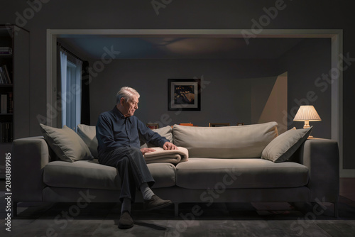 Elderly Man Sitting Alone on Large Sofa in Dimly Lit Living Room with Glowing Table Lamp, Senior Male at Home