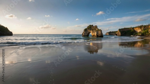 Serene coastal landscape featuring a natural rock arch formation with reflective wet sand and gentle ocean waves under a golden hour sky perfect for travel nature and relaxation concepts