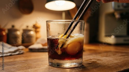 Raw egg yolk being stirred into dark beverage in glass with chopsticks on wooden kitchen countertop under warm ambient lighting for culinary concept