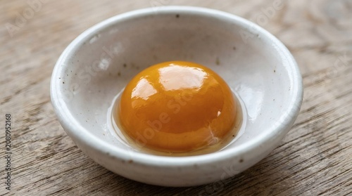 Fresh raw egg yolk centered in a small ceramic bowl on a rustic wooden tabletop surface for professional culinary ingredient photography