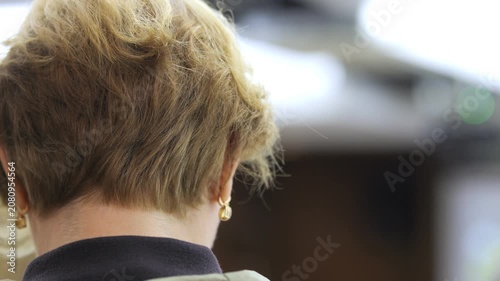 Close-up of woman's head, short blonde hair, earrings. She faces away, showing back of neck and collar. Soft focus background suggests indoor setting. Mood calm, focused, neutral