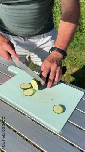 Close up of man hands slicing fresh green zucchini on plastic cutting board at campsite table.