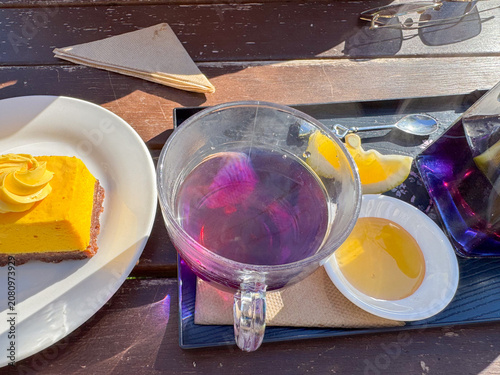 A glass cup of purple lavender tea served with lemon, honey, and a slice of bright yellow dessert on an outdoor café table. The colorful tea catches the sunlight, creating soft pink and violet reflect