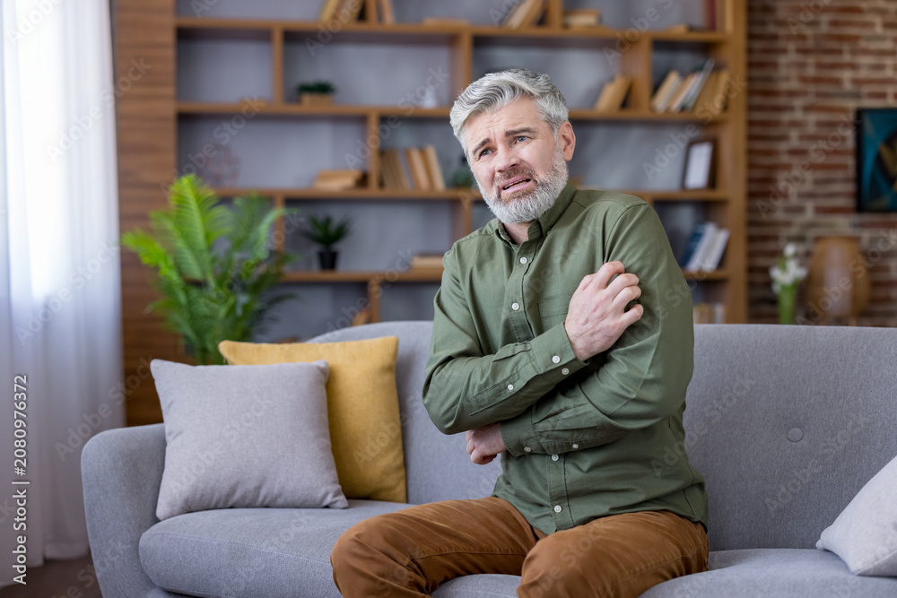 Fototapeta premium Mature man sitting uncomfortable on a sofa at home, scratching his arm and looking distressed, suffering from an allergic reaction or skin condition symptoms