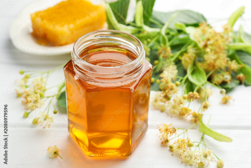 Naklejka premium Sweet honey in glass jar, honeycomb and linden flowers on white wooden table, closeup
