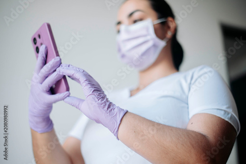 Low-angle view of a female nurse using a smartphone while wearing protective gloves and a face mask in a clinical setting.
