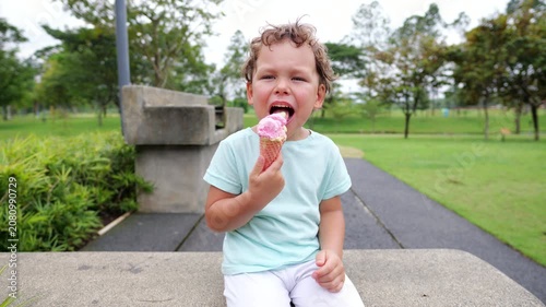 Happy little boy savoring a pink and yellow ice cream cone while sitting in green park, expressing pure joy and squinting slightly. Portrait shot with blurred background