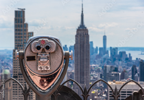 An iconic coin-operated observation telescope stands on a high-rise skyscraper deck, offering a scenic view of the Midtown Manhattan skyline with the prominent Empire State Building towering in the ba