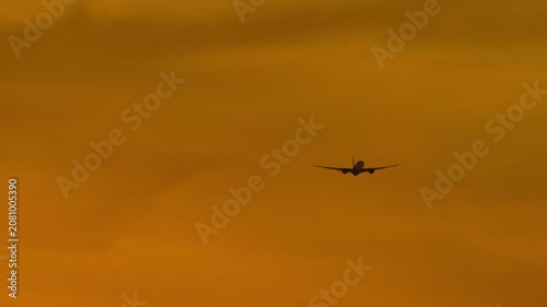 Commercial passenger airplane flying away in dramatic golden orange sunset sky with thick clouds, real-time telephoto silhouette shot