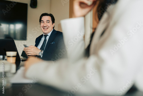 Smiling male business expert sitting with smart phone in meeting room at office