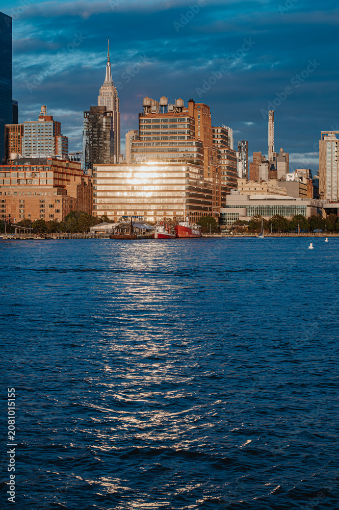 Fototapeta premium Waterfront buildings reflecting golden sunset light in Manhattan. Hudson River with ships docked at a pier and the Empire State Building behind.