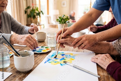 Elderly hands and caregiver hands work together on colorful watercolor painting at wooden table in warm care home room with cups, brushes, jars, and natural light for social creative activity
