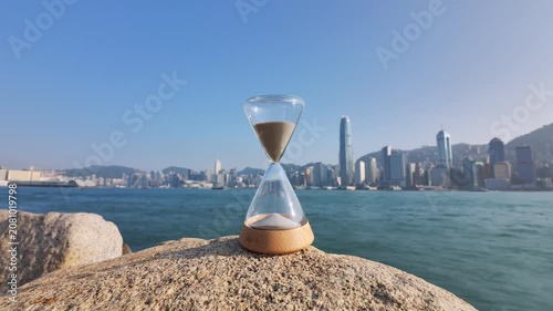 Time-lapse of hourglass standing at Hong Kong embankment with blurred Victoria Harbour and city waterfront on background. Conceptual shot of time's relentless passage with urban landscape backdrop